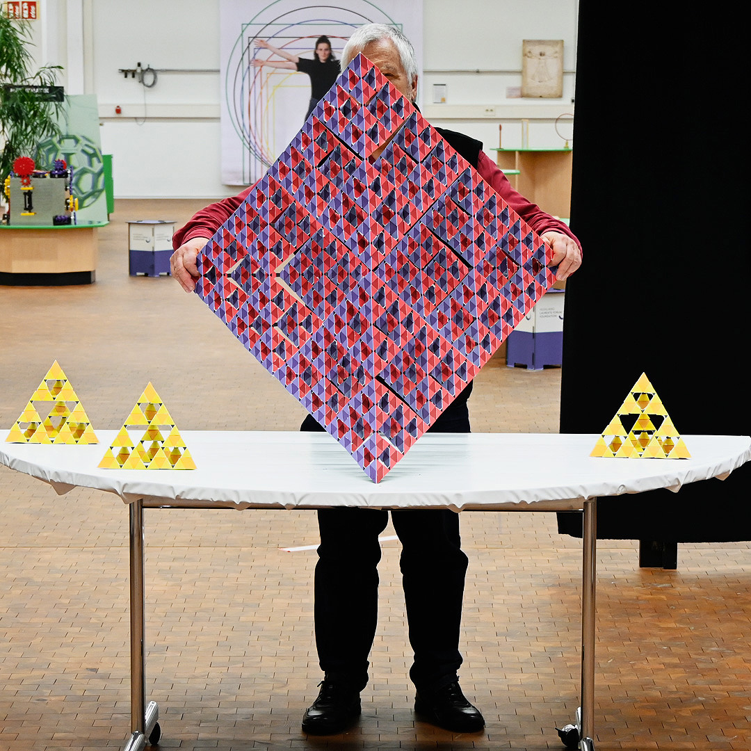 Interaktive Ausstellungsstation. Ein Mann mit grauen Haaren steht vor einem Tisch und hält einen großen platonischen Körper aus rotem und blauem Papier vor sich, sodass die Figur seinen gesamten Oberkörper verdeckt. 